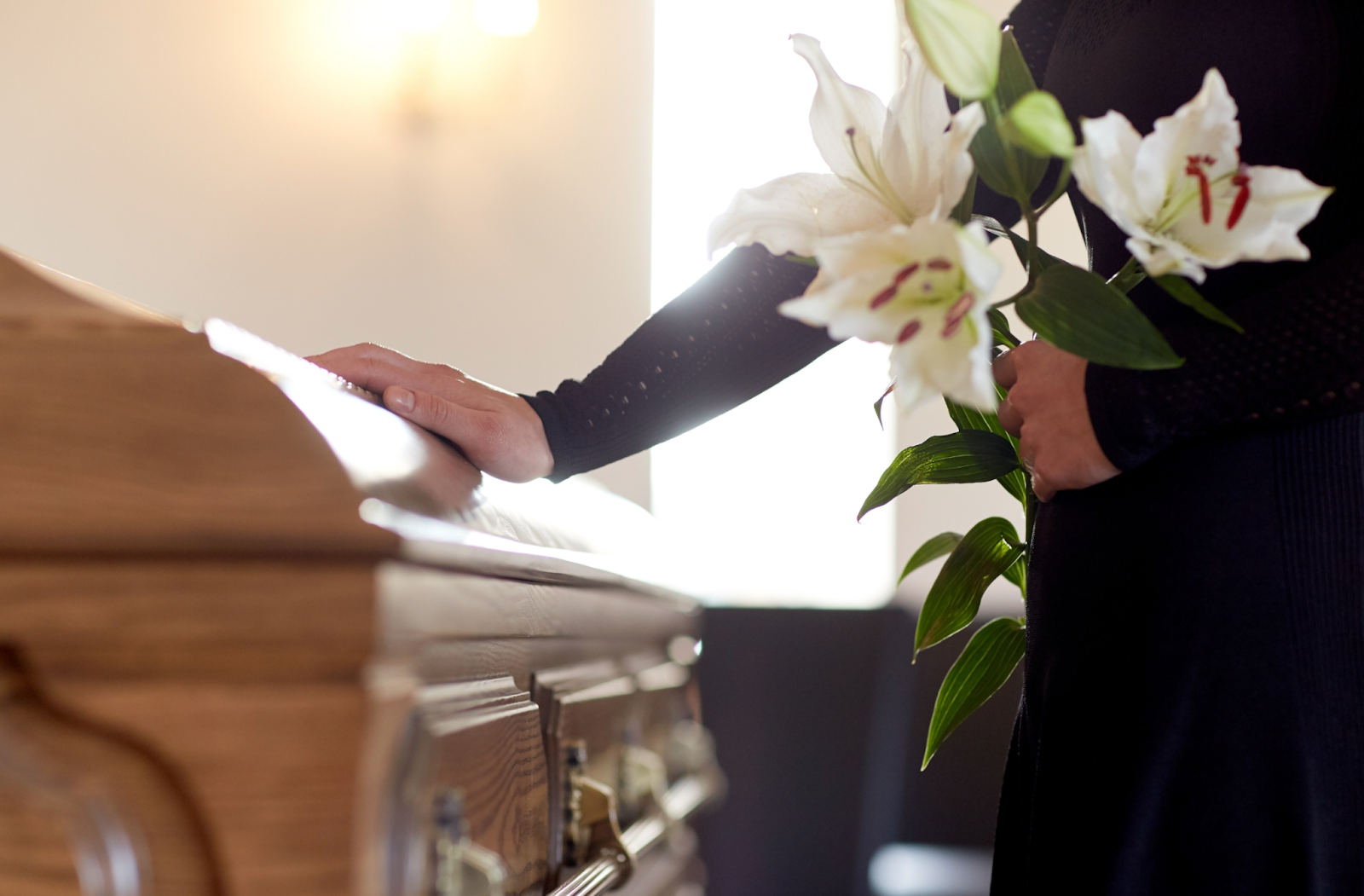 A person mourning at a funeral service, touching a casket with one hand and a bouquet of lilies in the other